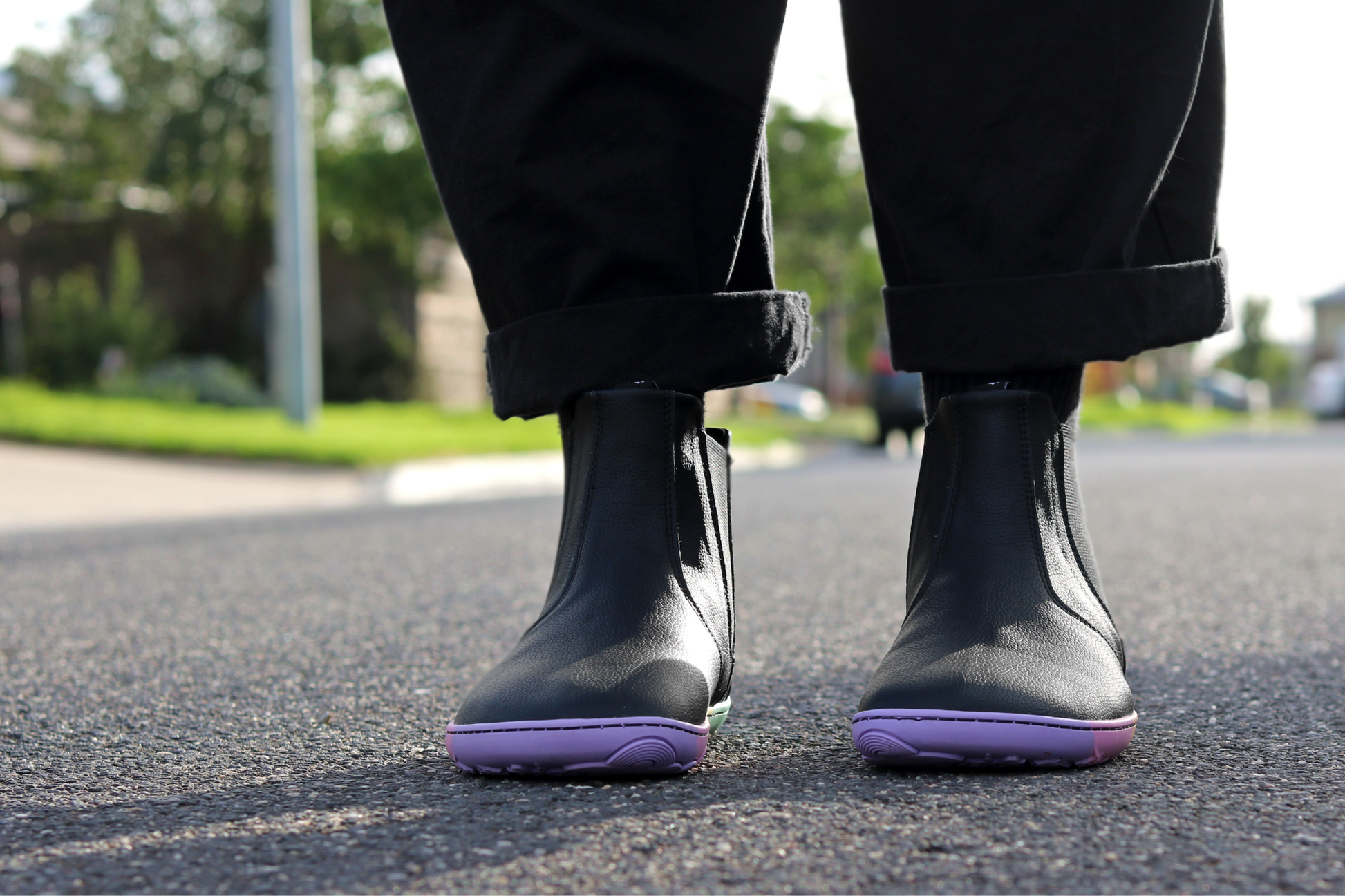 Black vegan leather chelsea boots from PaperKrane with rainbow foot-shaped sole on a street with grass in the background, the person is wearing black pants.