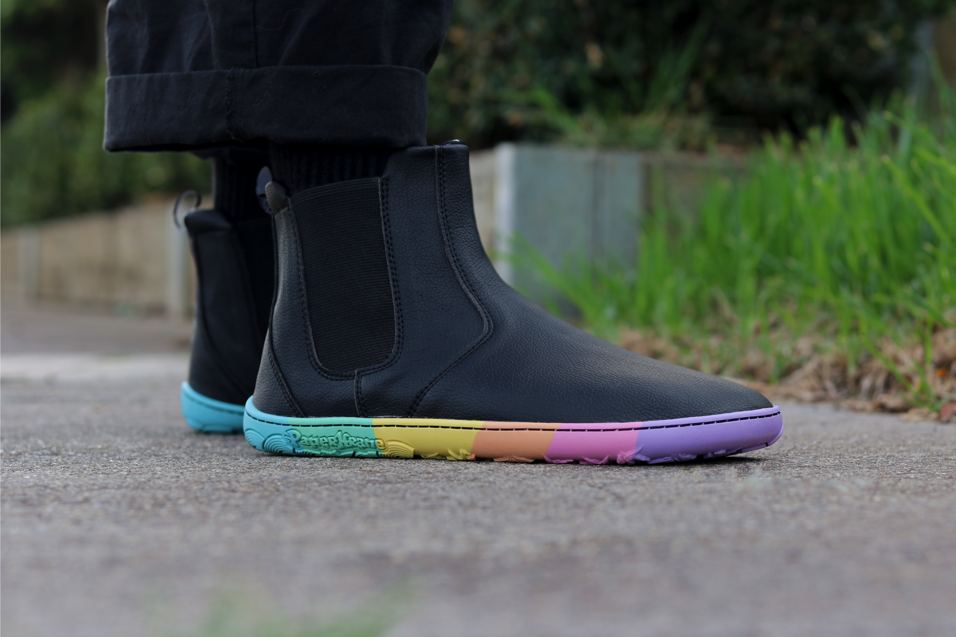 Black vegan leather chelsea boots from PaperKrane with rainbow foot-shaped sole on a footpath with grass in the background, the person is wearing black pants and socks, too. 
