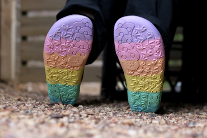  PaperKrane's rainbow foot-shaped soles, on a person who is sitting down. The ground is covered in stone chips. 