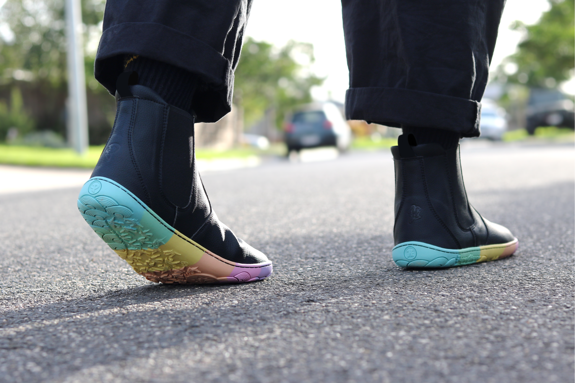 Black vegan leather chelsea boots from PaperKrane with rainbow foot-shaped sole on the street, a person is wearing black pants and socks and walking away in the boots.