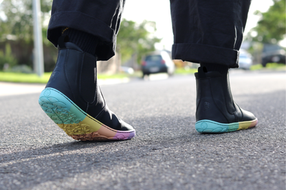Black vegan leather chelsea boots from PaperKrane with rainbow foot-shaped sole on the street, a person is wearing black pants and socks and walking away in the boots.