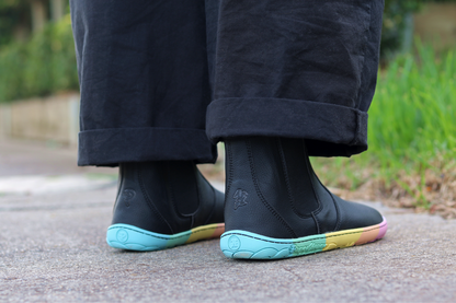 Back view of black vegan leather chelsea boots from PaperKrane with rainbow ZERO DROP sole on a footpath with grass in the background, the person is wearing black pants.