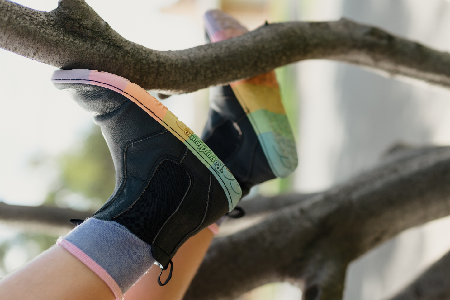 Black vegan leather chelsea boots from PaperKrane with rainbow foot-shaped sole climbing a tree. The child has purple socks on.
