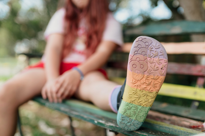 Black vegan leather chelsea boots from PaperKrane with rainbow foot-shaped sole on a bench. The sole is facing the camera and worn by a child. 