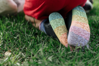 Black vegan leather chelsea boots from PaperKrane with rainbow foot-shaped soles. Worn by a child in red shorts, kneeling on grass. 