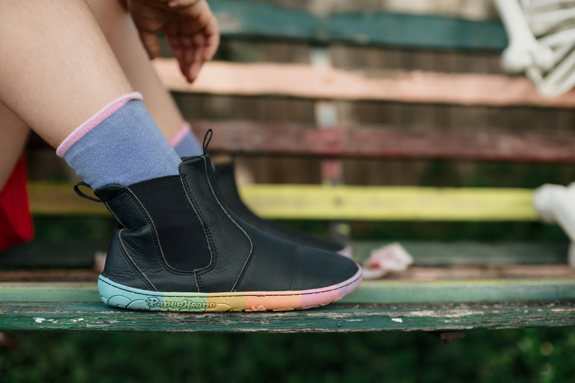 Black vegan leather chelsea boots from PaperKrane with rainbow zero drop sole on a bench. A child in purple socks is sitting on the bench in the flexible PaperKrane boots.