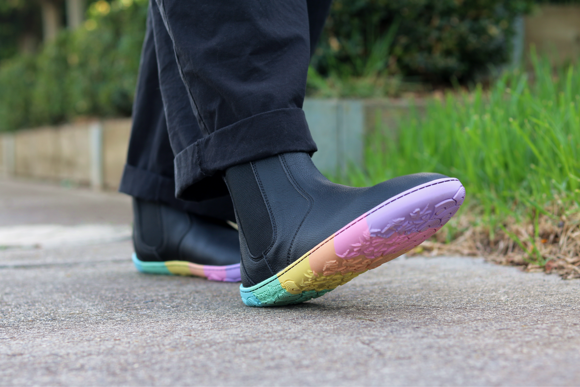 Black vegan leather chelsea boots from PaperKrane with rainbow foot-shaped soles walking on a footpath with grass in the background, the person is wearing black pants and socks, too.