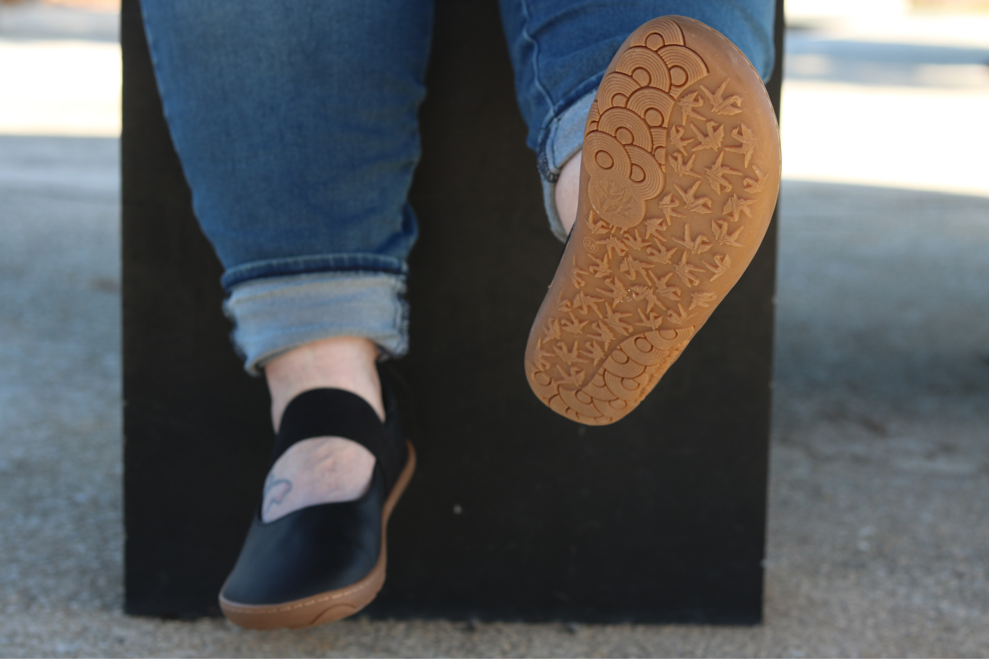 Adult feet in black vegan leather Licorice MJs, sitting on a bench with one foot raised to show the gum sole. 