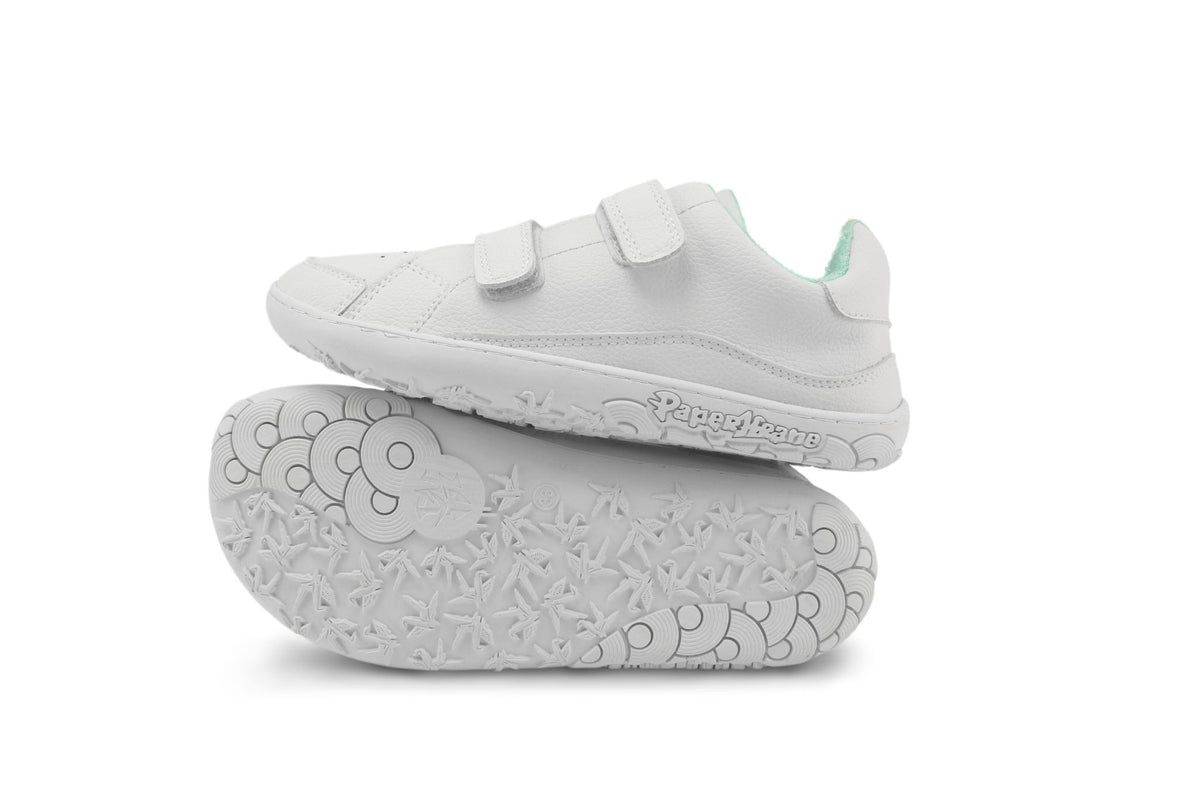 A pair of white kids' barefoot PaperKrane sneakers with velcro hook-and-loop straps, shown with one shoe standing upright and the other lying flat to display the textured sole, on a white background.