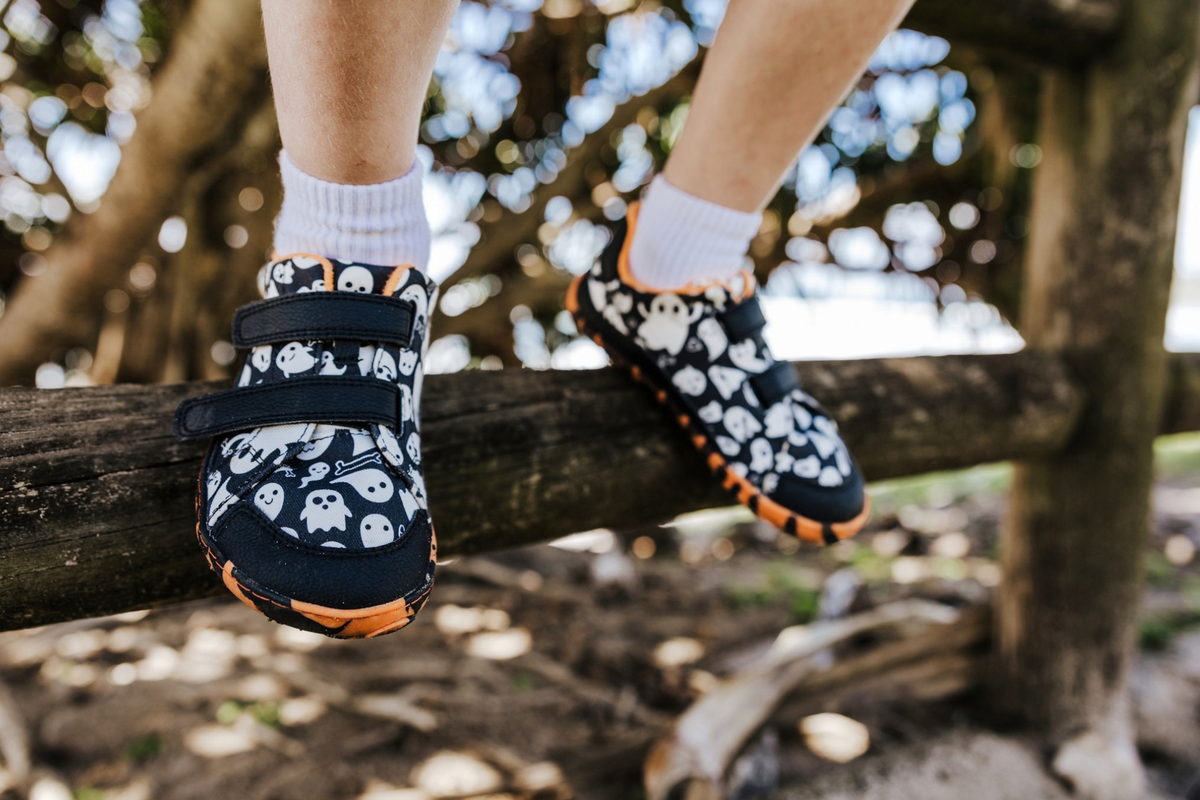 Orange striped barefoot soles with ghost print uppers with velcro straps are worn by a kid sitting on a fence. 