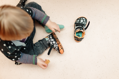 A small child sitting on a light beige floor wearing a pair of black and orange Halloween-themed PaperKrane velcro sneakers, with the matching bootie placed beside them.