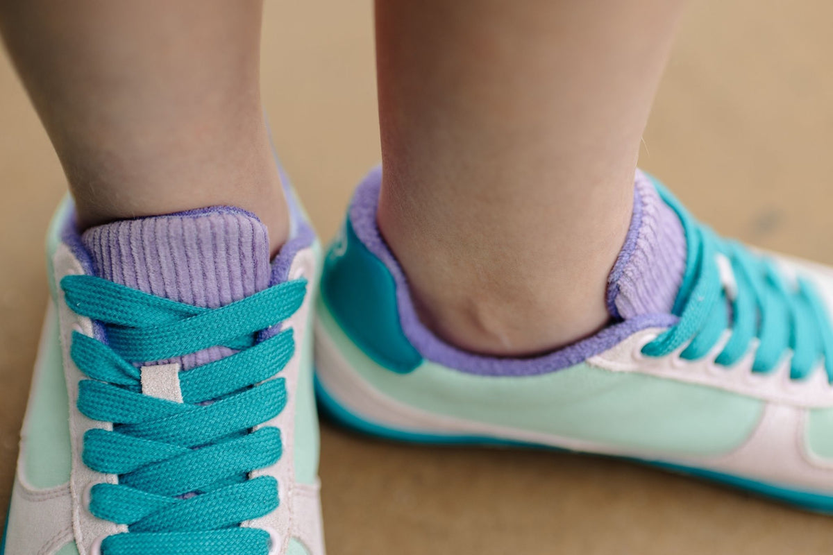 Close-up of a person's feet wearing pastel sneakers with teal laces, mint green panels, glittery pink leather details, soles, and lavender corduroy tongue, on a neutral concrete floor.