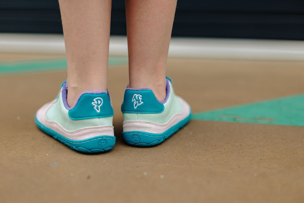 Close-up of a person's feet wearing mint green, glittery pink, purple and teal barefoot sneakers, with a P and K logo on the back, on a concrete surface.
