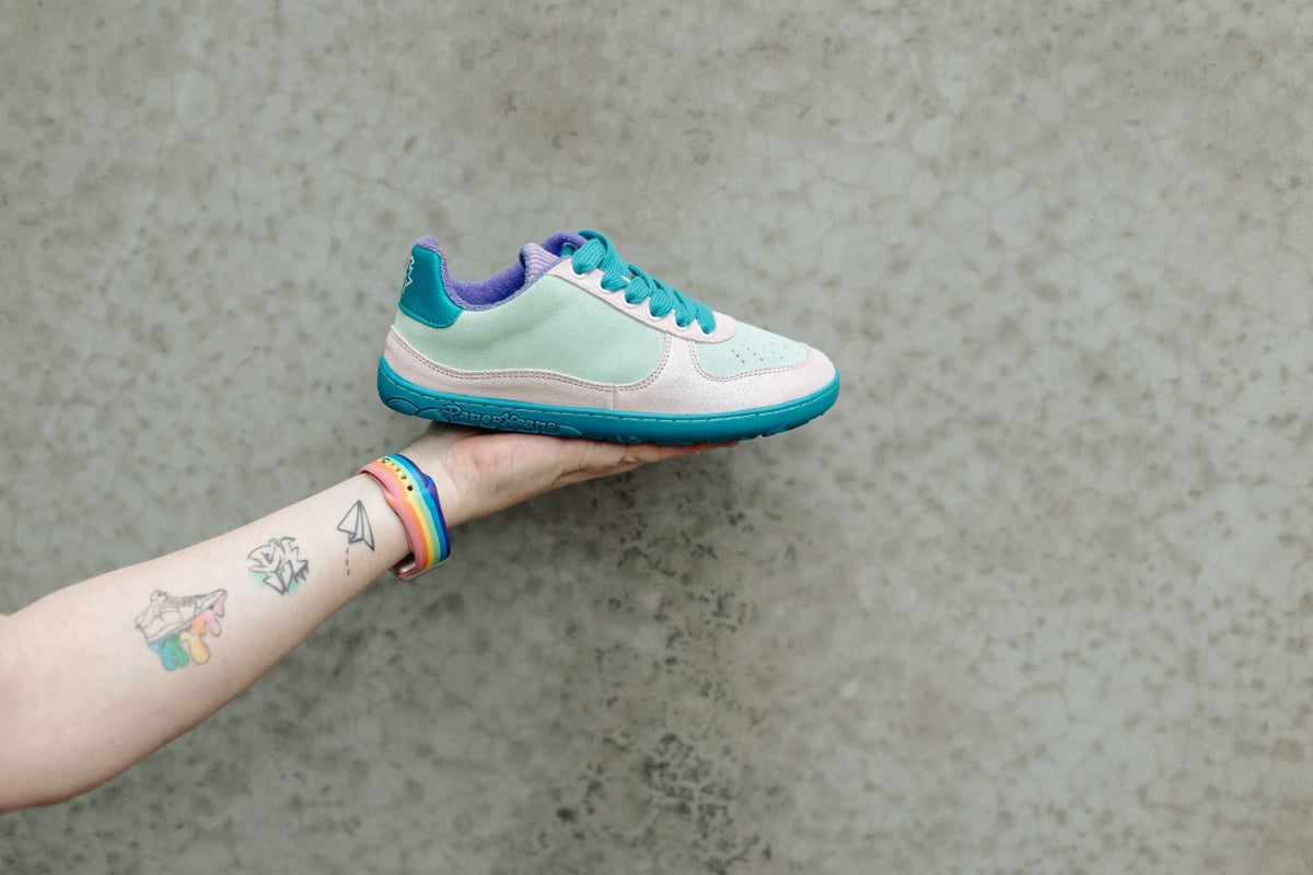 A mint green, glitter pastel pink, and teal low-top barefoot sneaker held in a person's hand against a textured grey concrete background.