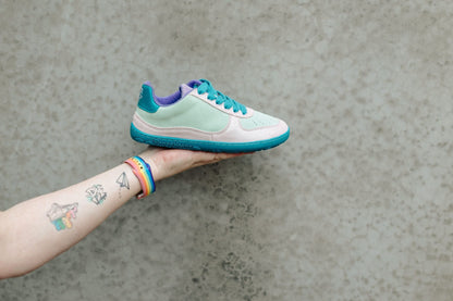 A mint green, glitter pastel pink, and teal low-top barefoot sneaker held in a person's hand against a textured grey concrete background.