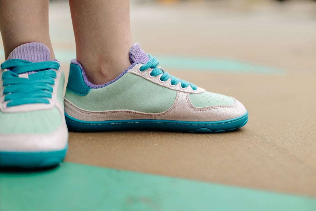 A close-up of a person's feet wearing pastel mint green, glittery pink, lavender, and teal low-top barefoot sneakers on a concrete outdoor surface.
