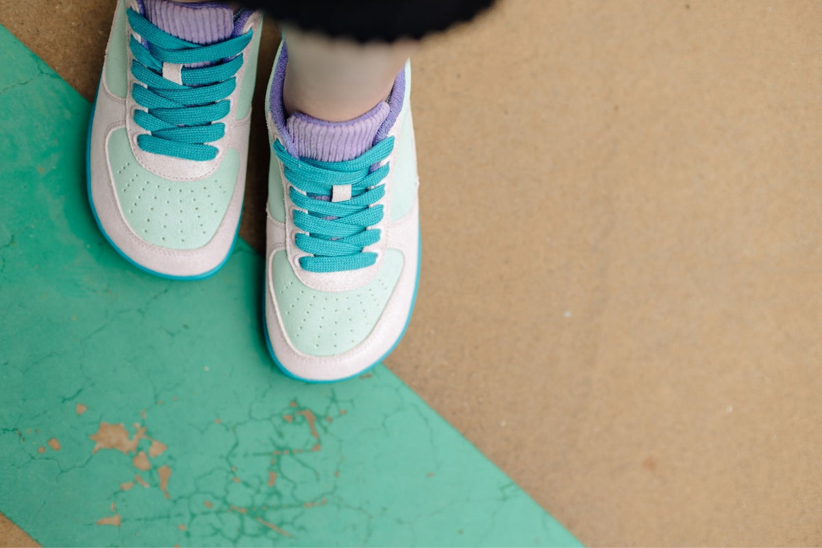 A close-up, top-down view of a person's feet wearing mint green and glittery pastel pink foot-shaped sneakers with bright teal laces and teal rubber sole, standing on a painted concrete surface.