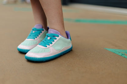 A close-up shot of a person's feet wearing pastel mint green, pale glittery pink, and teal sneakers with purple corduroy tongue and terry lining, standing on a brown concrete surface.
