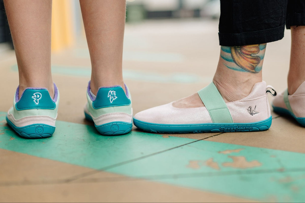 Close-up of two pairs of flat shoes worn by people standing outdoors on a painted concrete surface. The shoes are pastel glittery pink with mint green details and teal soles, the mary jane style has small embossed krane logos on the sides.