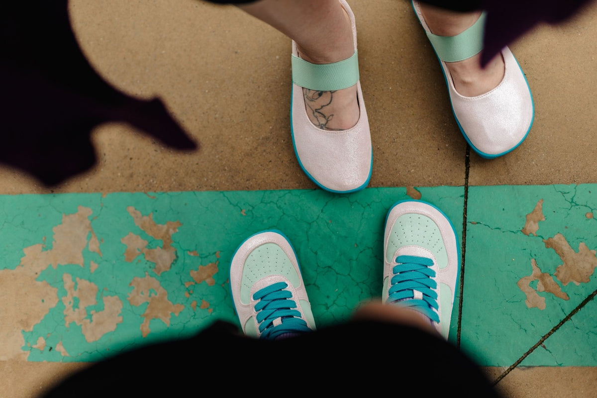 A top-down view of two pairs of shoes on feet. The shoes are pale glittery pink with mint green and bright teal accents, including laces and soles, shown standing on a painted concrete floor.