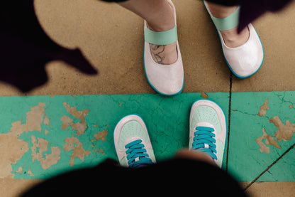 A top-down view of two pairs of shoes on feet. The shoes are pale glittery pink with mint green and bright teal accents, including laces and soles, shown standing on a painted concrete floor.