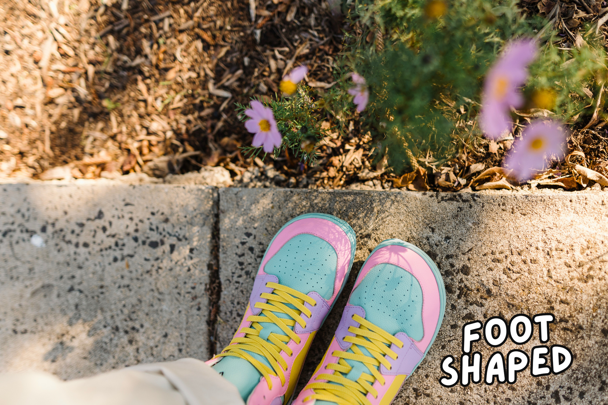 Pastel-colored shoes with yellow laces on a concrete surface with flowers in the background.