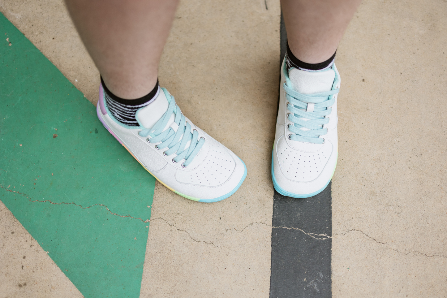 White sneakers on rainbow barefoot soles, with light blue accents worn on a concrete surface.