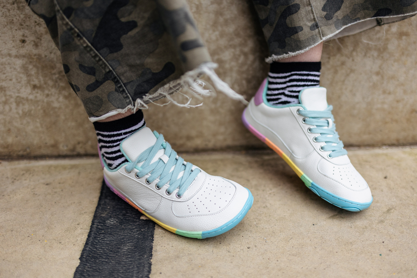 White sneakers with rainbow soles and light blue laces worn with camouflage pants and striped socks.