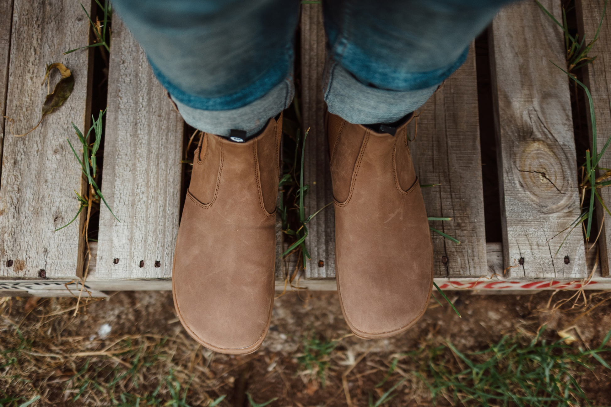 Brown leather foot-shaped chelsea boots from PaperKrane and blue jeans on a wooden platform with grass and dirt below