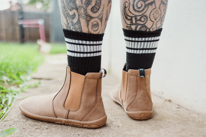 Brown leather PaperKrane barefoot chelsea boots worn with black and white striped socks on a concrete surface. The person has tattooed legs.
