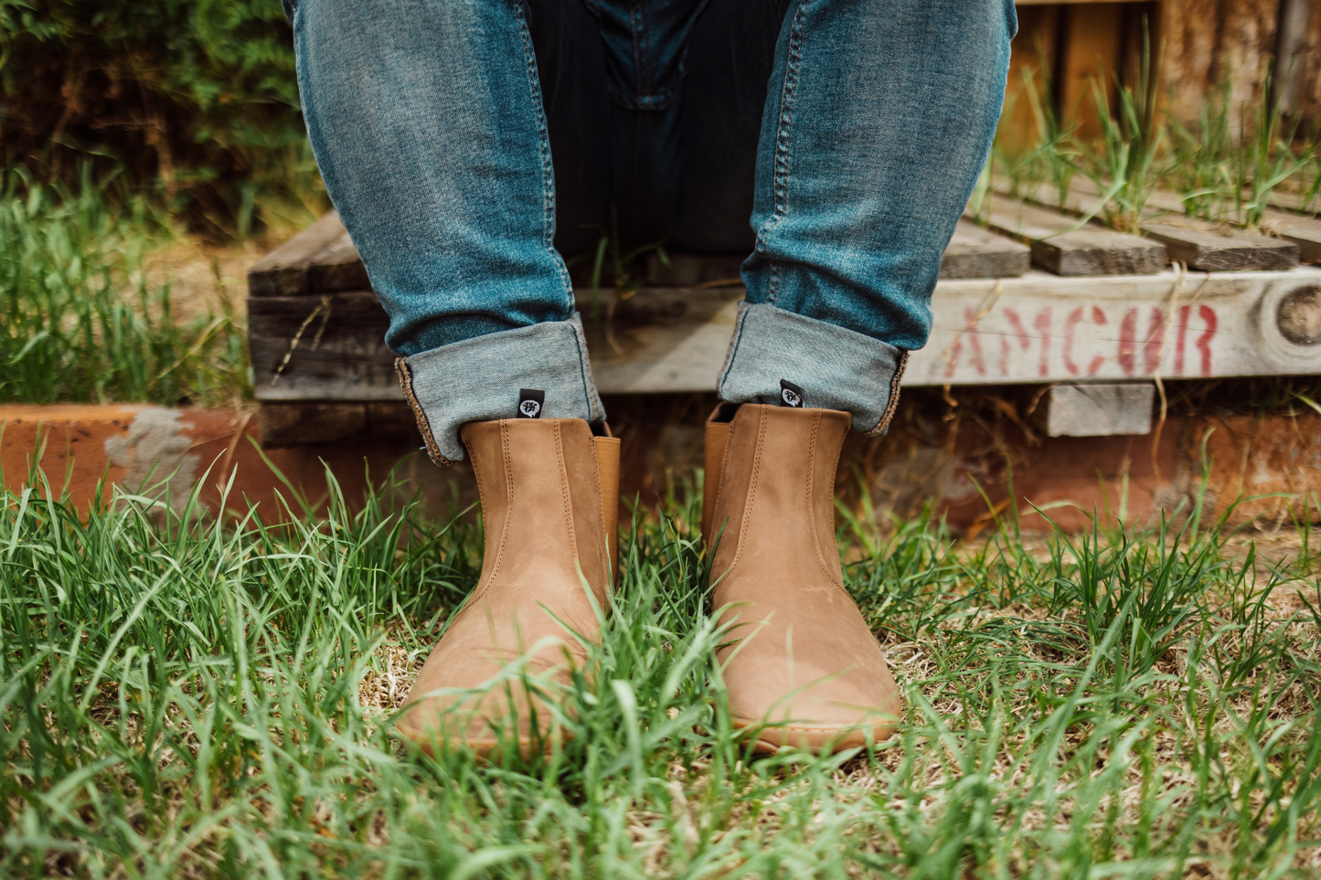 Person wearing brown leather Paperkrane barefoot boots and rolled-up jeans sitting on a pallet with feet on grass with a blurred background