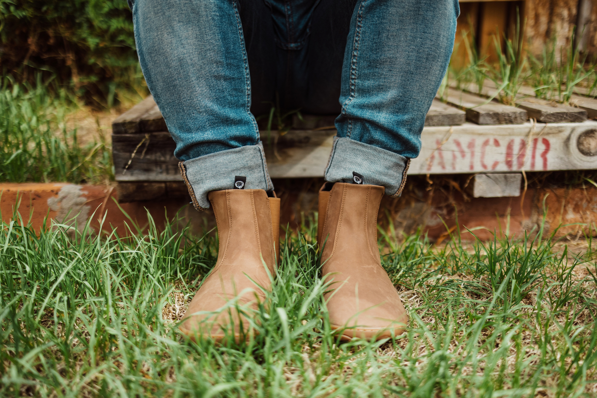 Person wearing brown leather Paperkrane barefoot boots and rolled-up jeans sitting on a pallet with feet on grass with a blurred background