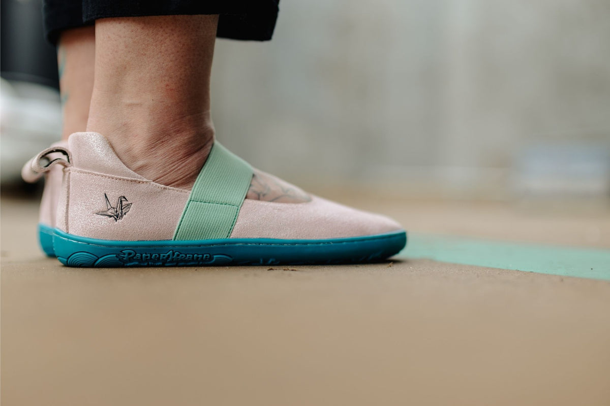 A close-up of a person's foot wearing a PaperKrane mary jane slip-on shoe. The shoe features a light shimmery pink leather upper, a mint green elastic strap, and a bright teal-blue sole, shown standing on a concrete floor.