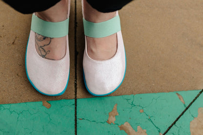 A close-up of a person's feet wearing light pink leather Mary Jane-style shoes with a glittery appearance, mint green elastic straps and bright teal soles, standing on a concrete floor with a green painted section below.