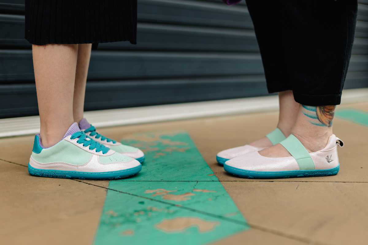 A close-up lifestyle shot of two people standing on a concrete ground, showcasing a pair of pastel mint green and turquoise barefoot sneakers on one side and a pair of matchy pastel pink leather mjs, with teal soles and mint elastic straps on the other.
