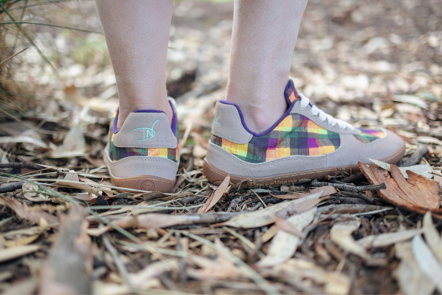 View from the back of a pair of tan PaperKrane barefoot sneakers with Di Nowinski plaid pattern on some dried leaves and branches of the Australian bush. 