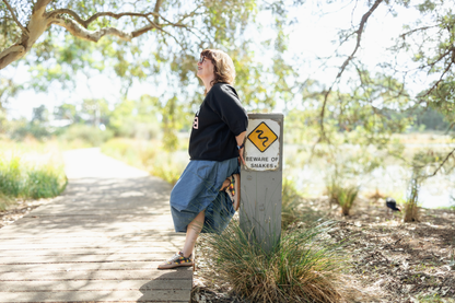 Artist Di Nowinski standing on a wooden path next to a 'Beware of Snakes' sign, she is wearing PaperKrane barefoot sneakers with her own plaid art print on them, with trees and nature in the background.