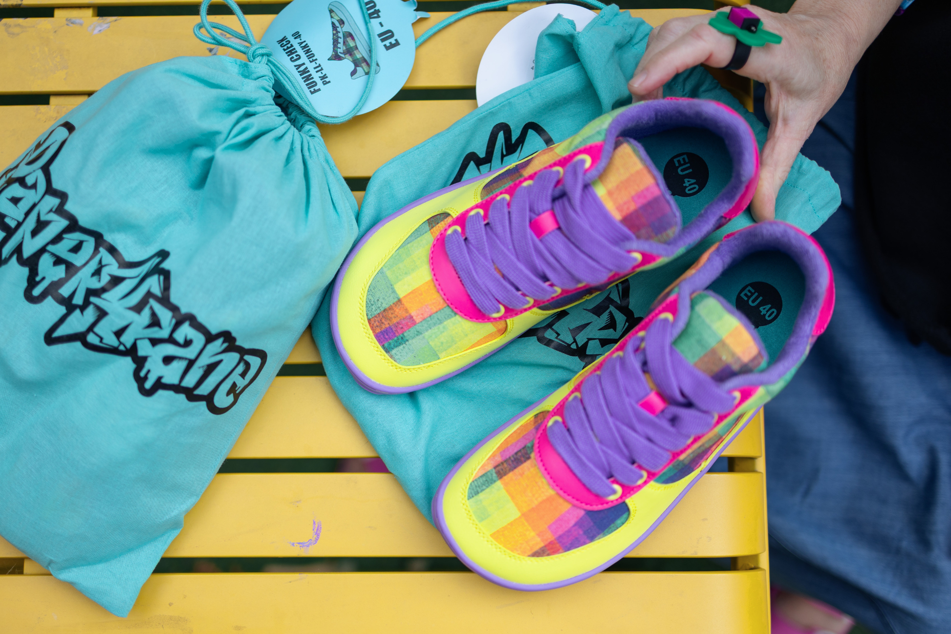 Colourful barefoot foot-shaped PaperKrane sneakers with a teal drawstring bag on a yellow wooden surface. The colourful shoes feature an art print by Artist, Di Nowinski. 