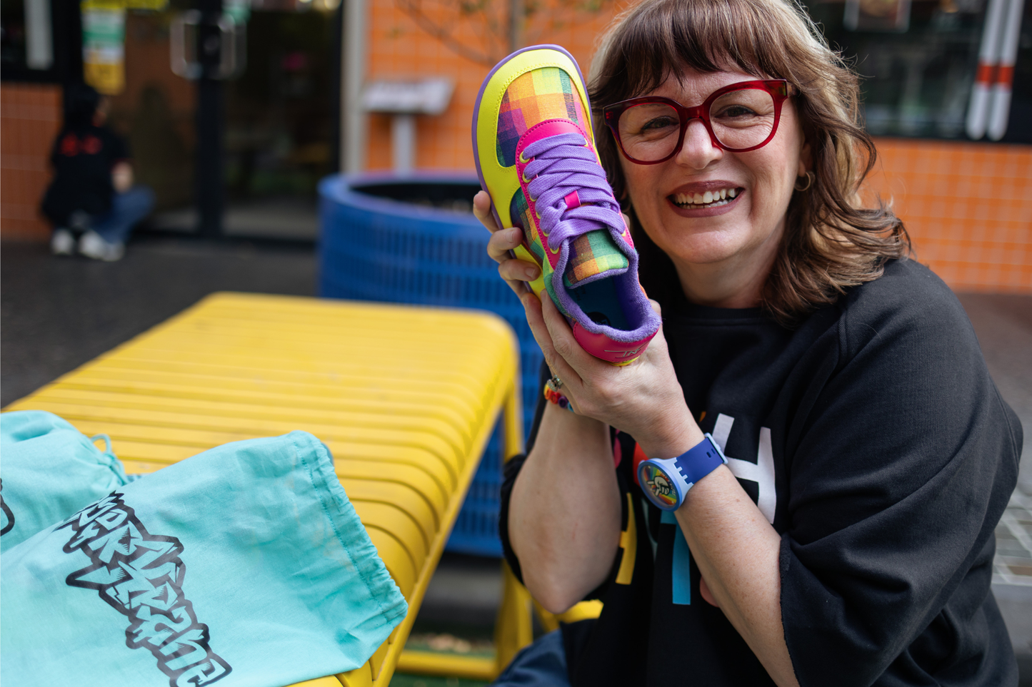 Di Nowinski holding one Checkered Joy shoe and smiling. The shoes are yellow, pink, purple and feature a plaid print by Di. They're a foot-shaped collab with barefoot shoe brand, PaperKrane