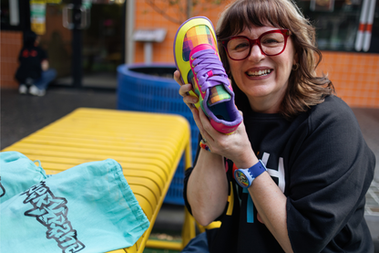 Di Nowinski holding one Checkered Joy shoe and smiling. The shoes are yellow, pink, purple and feature a plaid print by Di. They're a foot-shaped collab with barefoot shoe brand, PaperKrane
