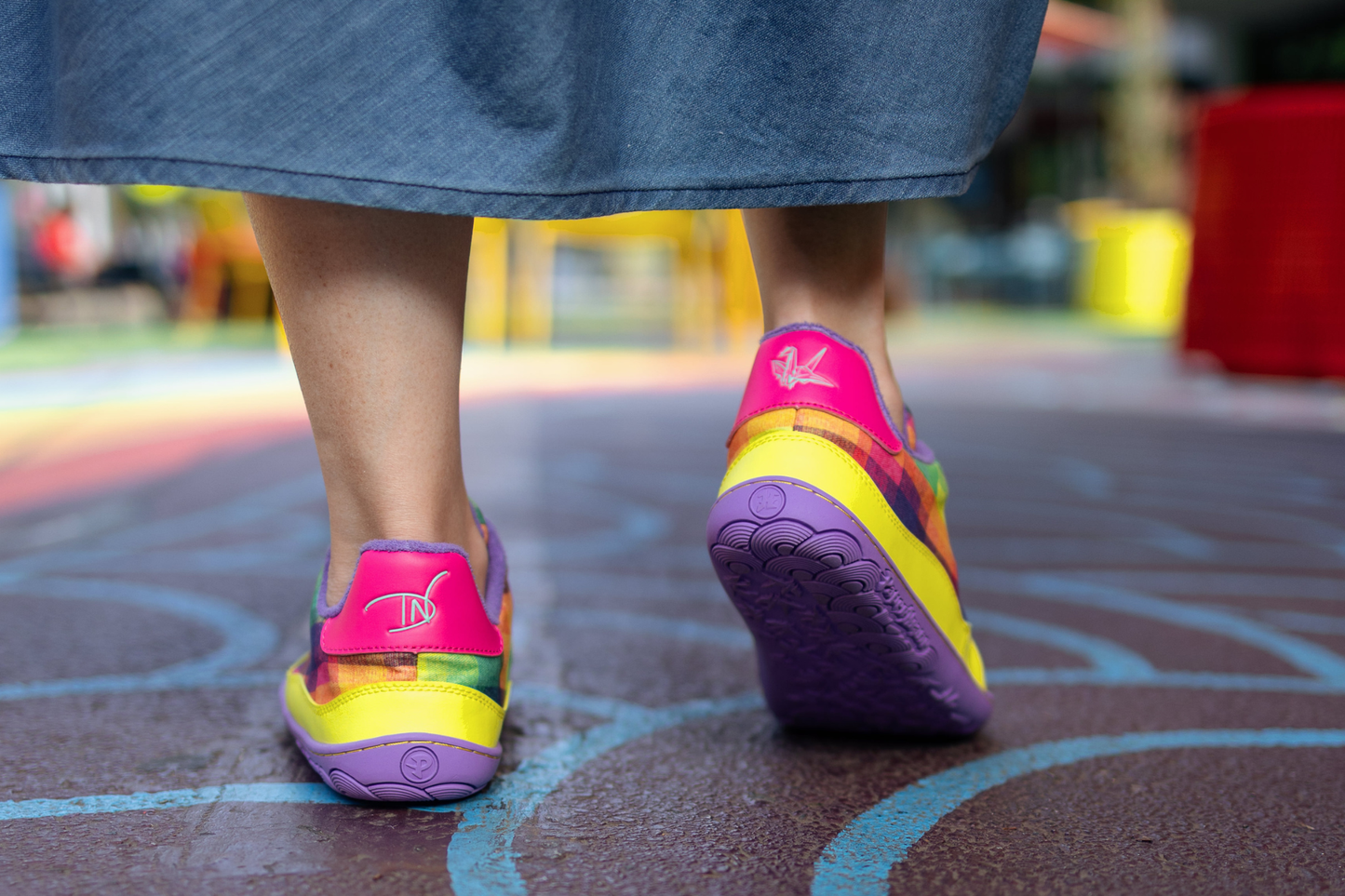Colourful PaperKrane x Di Nowinski collab shoes (with plaid print, pink, yellow, and purple details) on a playground surface with a blurred background