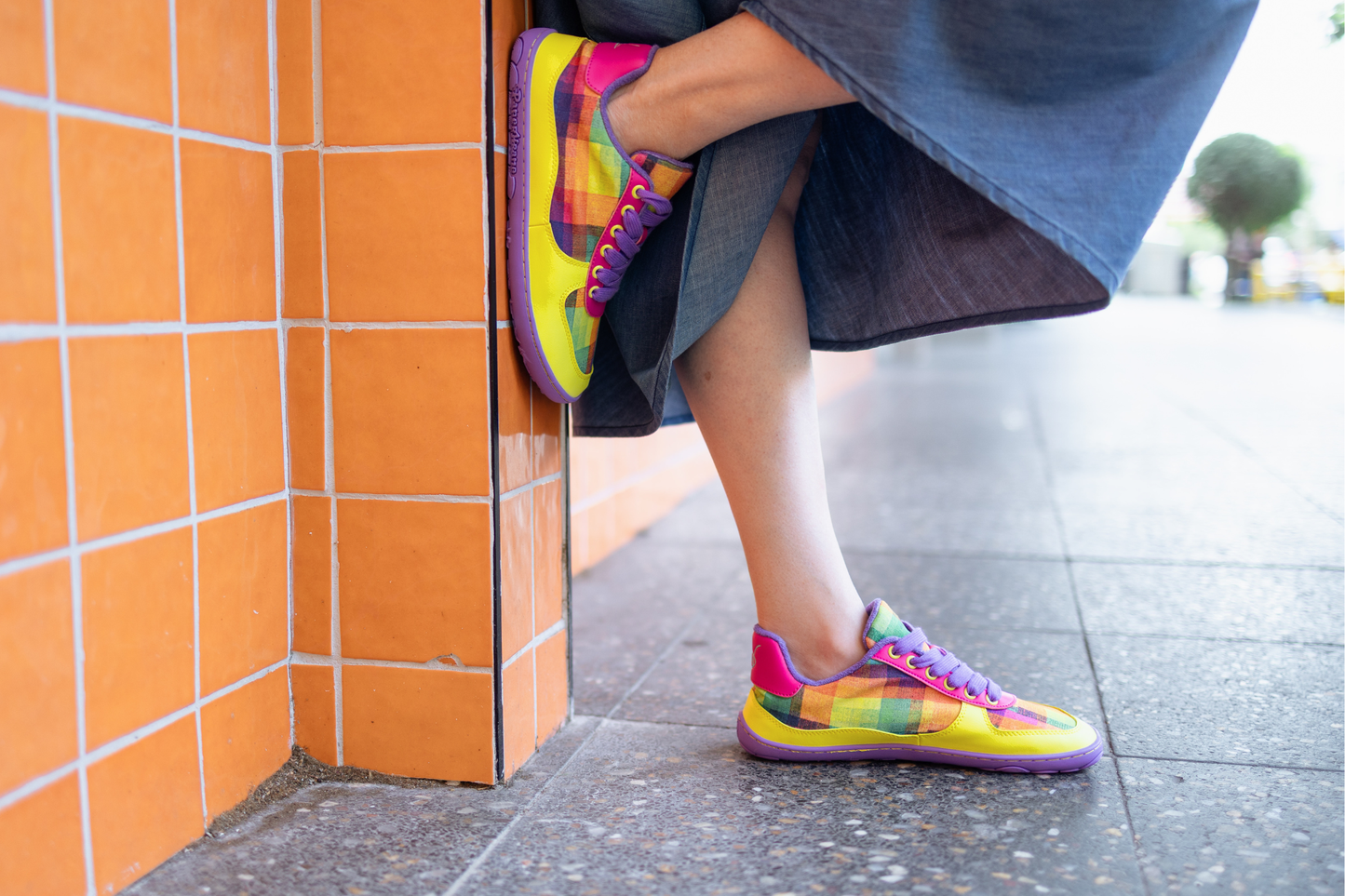 Person wears a denim skirt and leans against and orange wall with bright barefoot PaperKrane sneakers. Featuring Di Nowinski plaid print. 