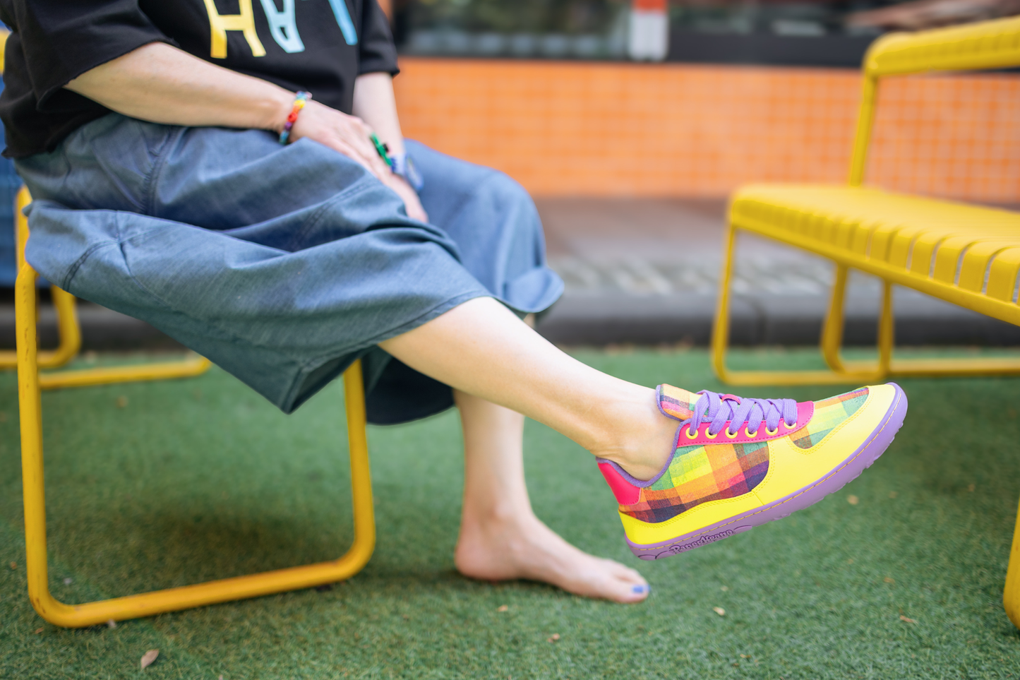 Person sitting on a yellow bench wearing colourful PaperKrane x Di Nowinski collab barefoot sneakers with a blurred background