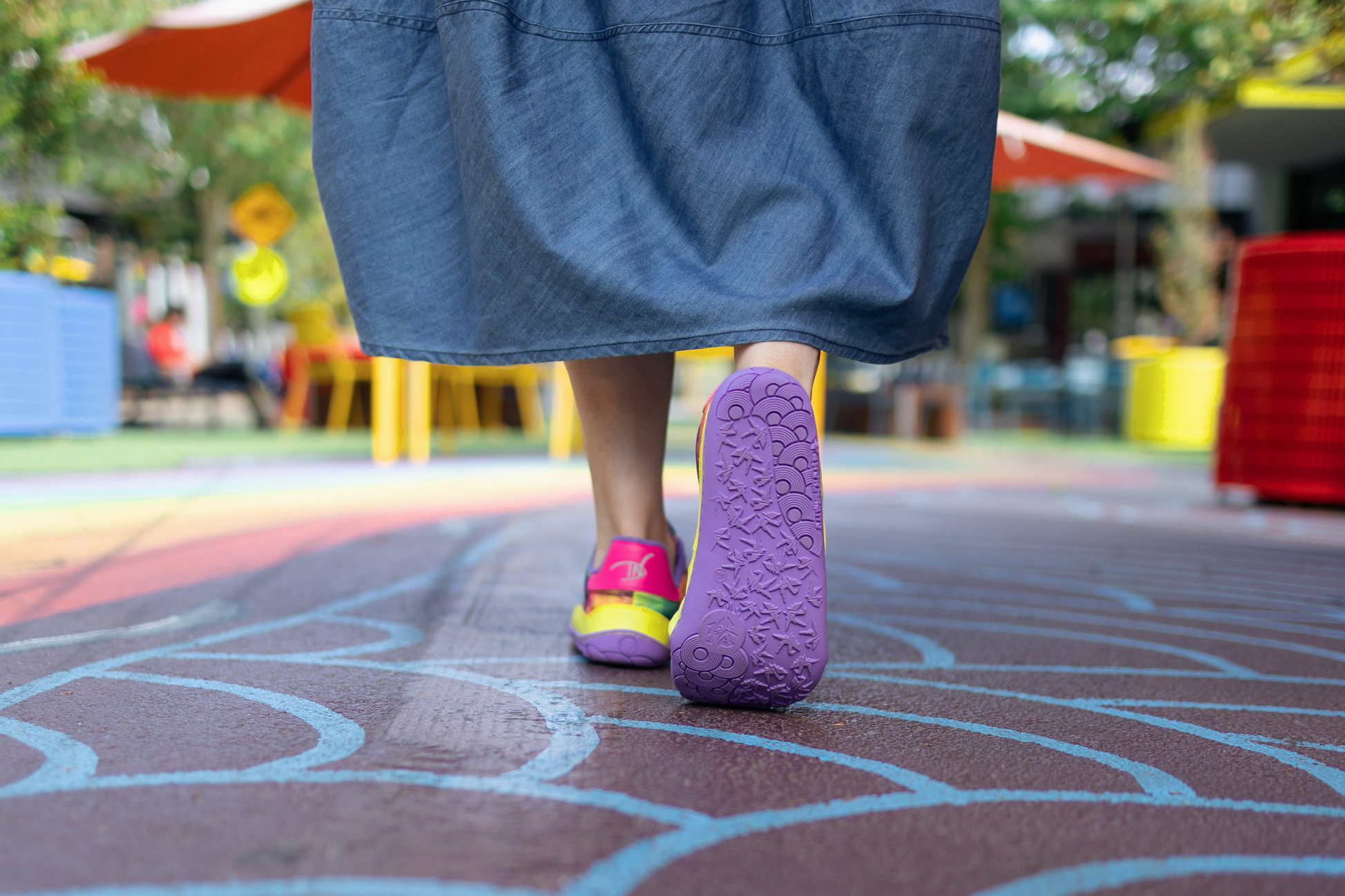 Di Nowinski wearing a blue skirt and colorful barefoot PaperKrane shoes with a purple sole, on a patterned ground with blurred background