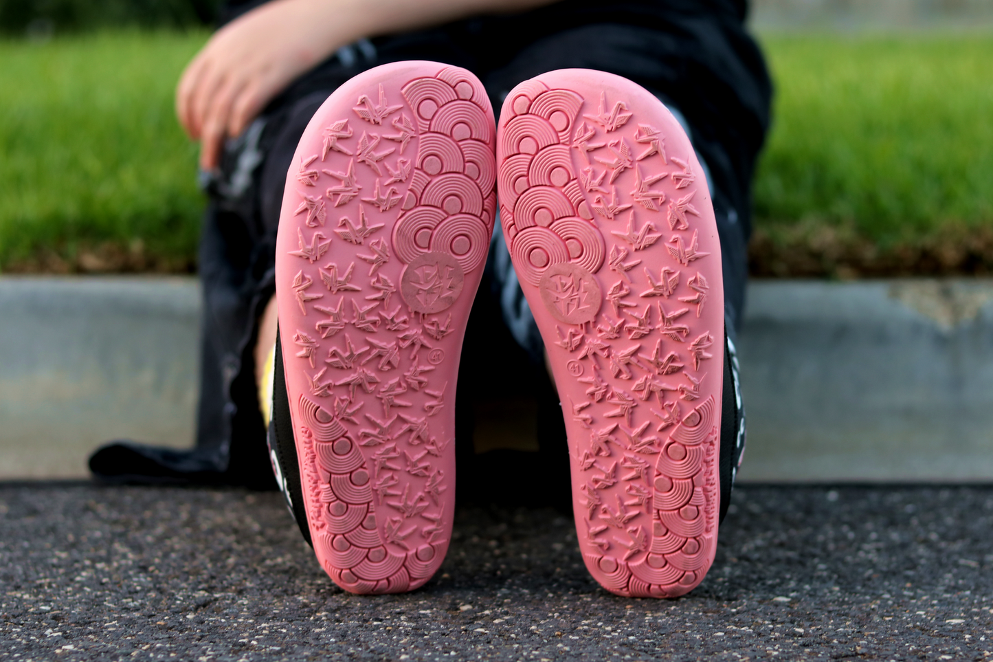 The salmon rubber sole of a pair of PaperKrane barefoot eyeball boots on a street. The boots are black with cartoon eyeballs, elastic laces, they're a foot-shaped, zero-drop design, from this angle we can see the full shape of the soles and the krane + rainbow grip design on the bottom. 