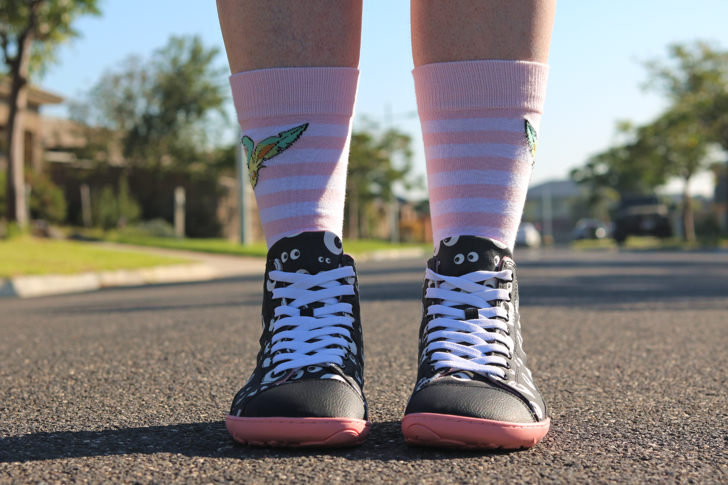 PaperKrane barefoot eyeball boots on legs with stripey socks that are facing towards the camera, standing on a street. The boots are black with cartoon eyeballs, elastic laces, they're a foot-shaped, zero-drop design, on a salmon coloured sole. 