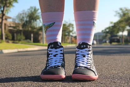 PaperKrane barefoot eyeball boots on legs with stripey socks that are facing towards the camera, standing on a street. The boots are black with cartoon eyeballs, elastic laces, they're a foot-shaped, zero-drop design, on a salmon coloured sole. 