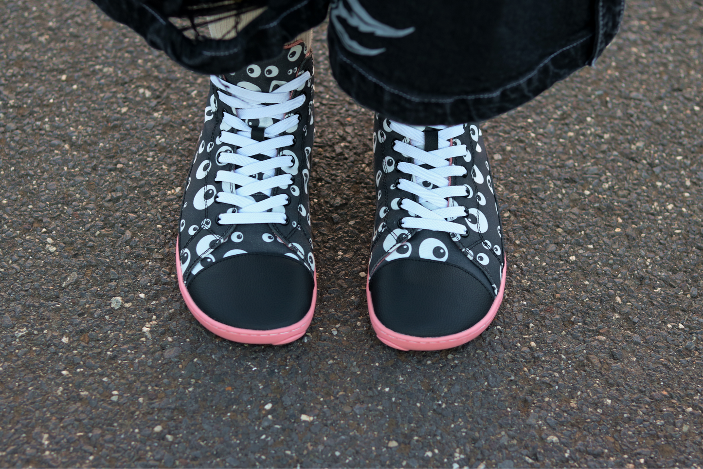 Above view of PaperKrane barefoot eyeball boots on a young person with some ripped shorts, on a street. The boots are black with cartoon eyeballs, elastic laces, they're a foot-shaped, zero-drop design, on a salmon coloured sole. 