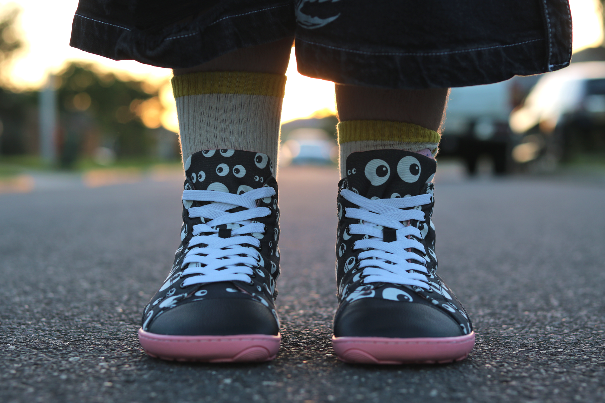 Front view of PaperKrane barefoot eyeball boots on a young person's legs with funky socks and ripped shorts, on a street. The boots are black with cartoon eyeballs, elastic laces, they're a foot-shaped, zero-drop design, on a salmon coloured sole. 