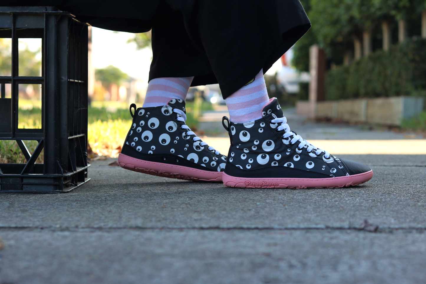 PaperKrane barefoot eyeball boots on legs with stripey socks, on a footpath. The person is sitting on a crate wearing boots that are black with cartoon eyeballs, elastic laces, they're a foot-shaped, zero-drop design, on a salmon coloured sole. 